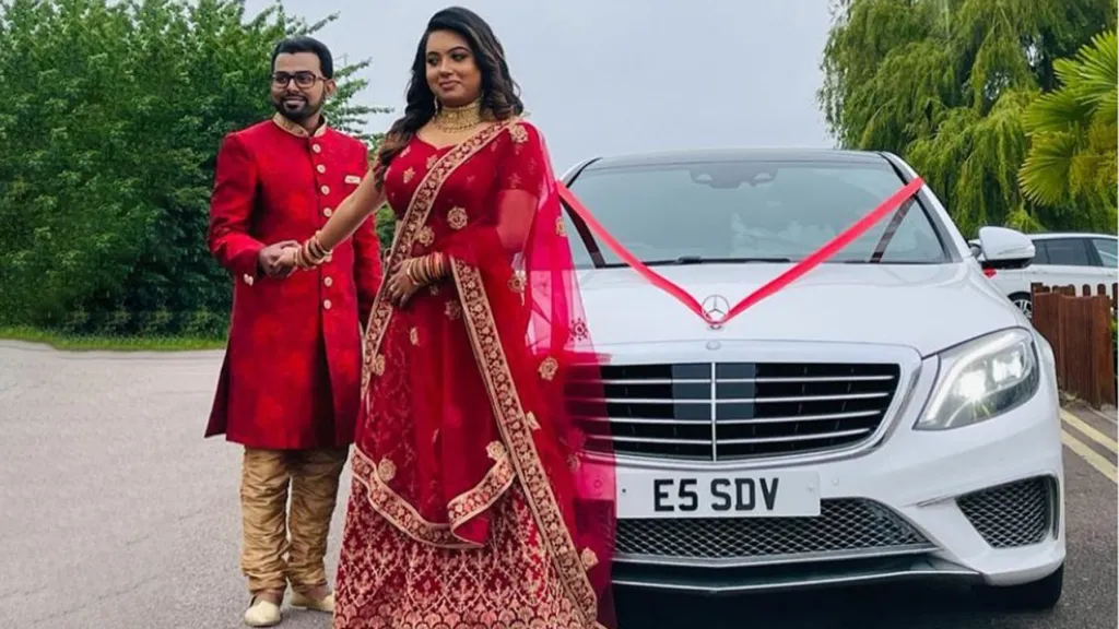 White Mercedes S-Class decorated with Red Ribbons accros its bonnet. Both Bride and Groom wearing traditional Asian Wedding Gowns are standing in front of the vehicle holding hands.