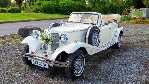 A Vintage Beauford in Ivory with convertible roof open showing a cream leather interior. Car is decorated with floral bouquet on its front radiator. Chrome spokes wheels with thin whitewall tires and two spare wheels mounted on both side of the car.