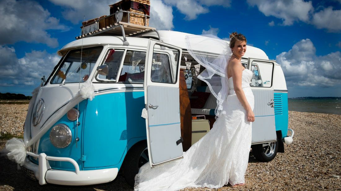 Smiling Bride in her white dress standing in front of a Blue & White Classic VW campervan decorated with white wedding ribbons