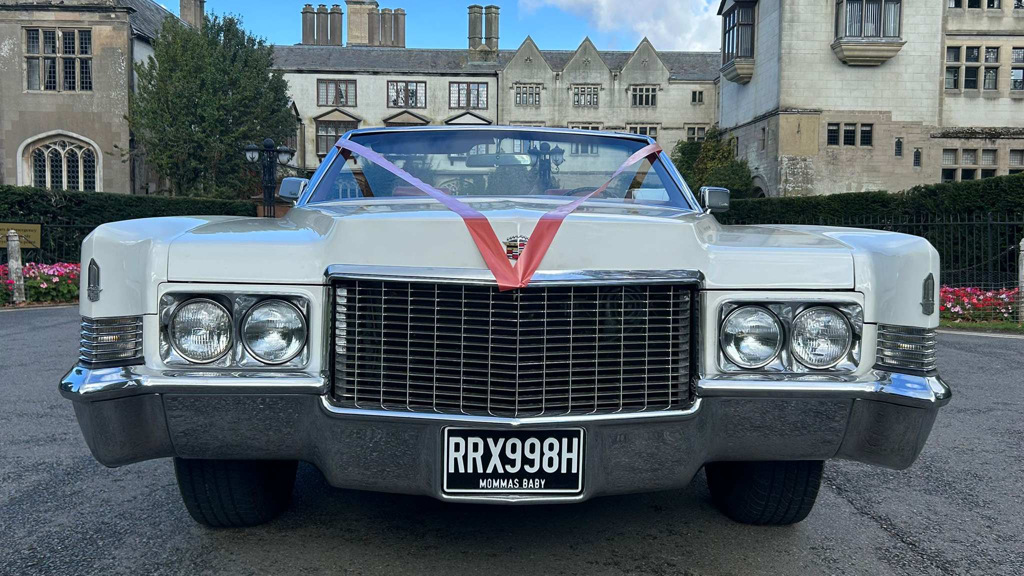 Close-up front view of a white 1970 Cadillac Coupe DeVille showing its distinctive chrome grille, quad headlights, and red wedding ribbons.