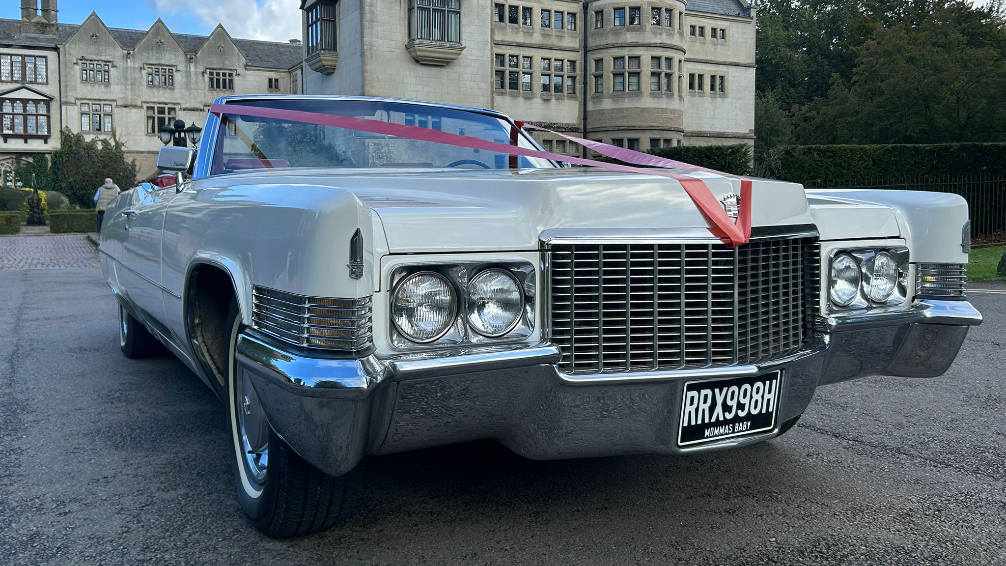 White 1970 Cadillac Coupe DeVille convertible decorated with white wedding ribbons, parked in front of an elegant manor venue and decorated with red coloured wedding ribbons