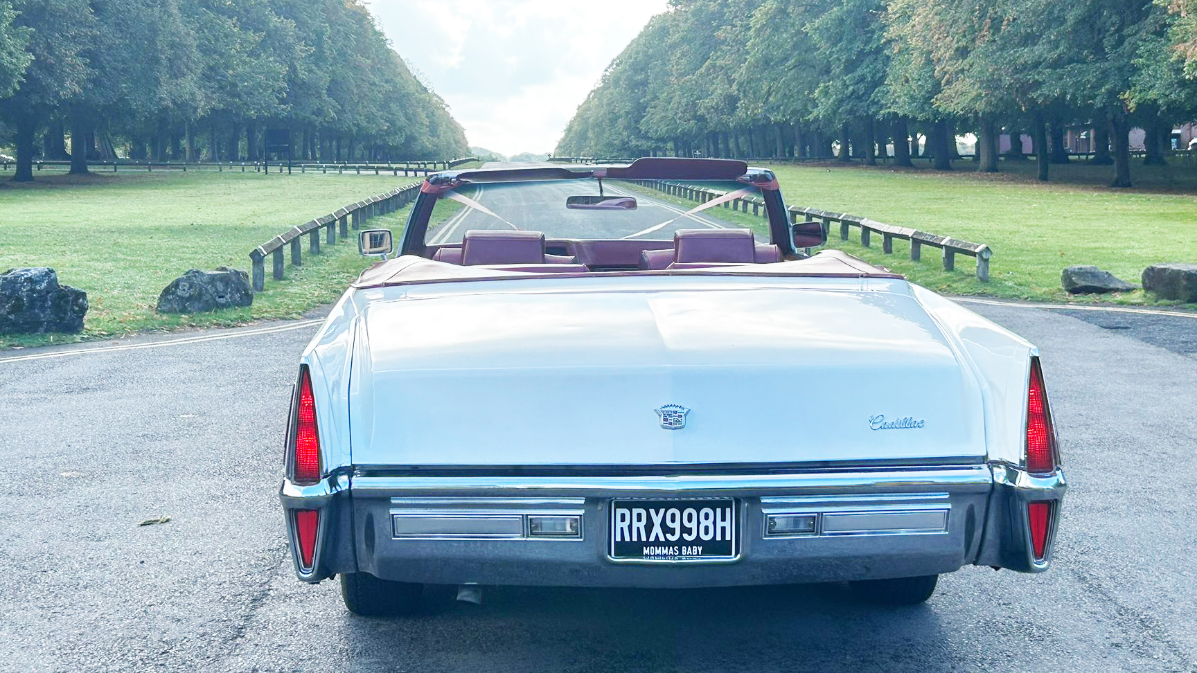 Rear view of a white 1970 Cadillac Coupe DeVille convertible with red interior, parked on a wide tree-lined driveway.