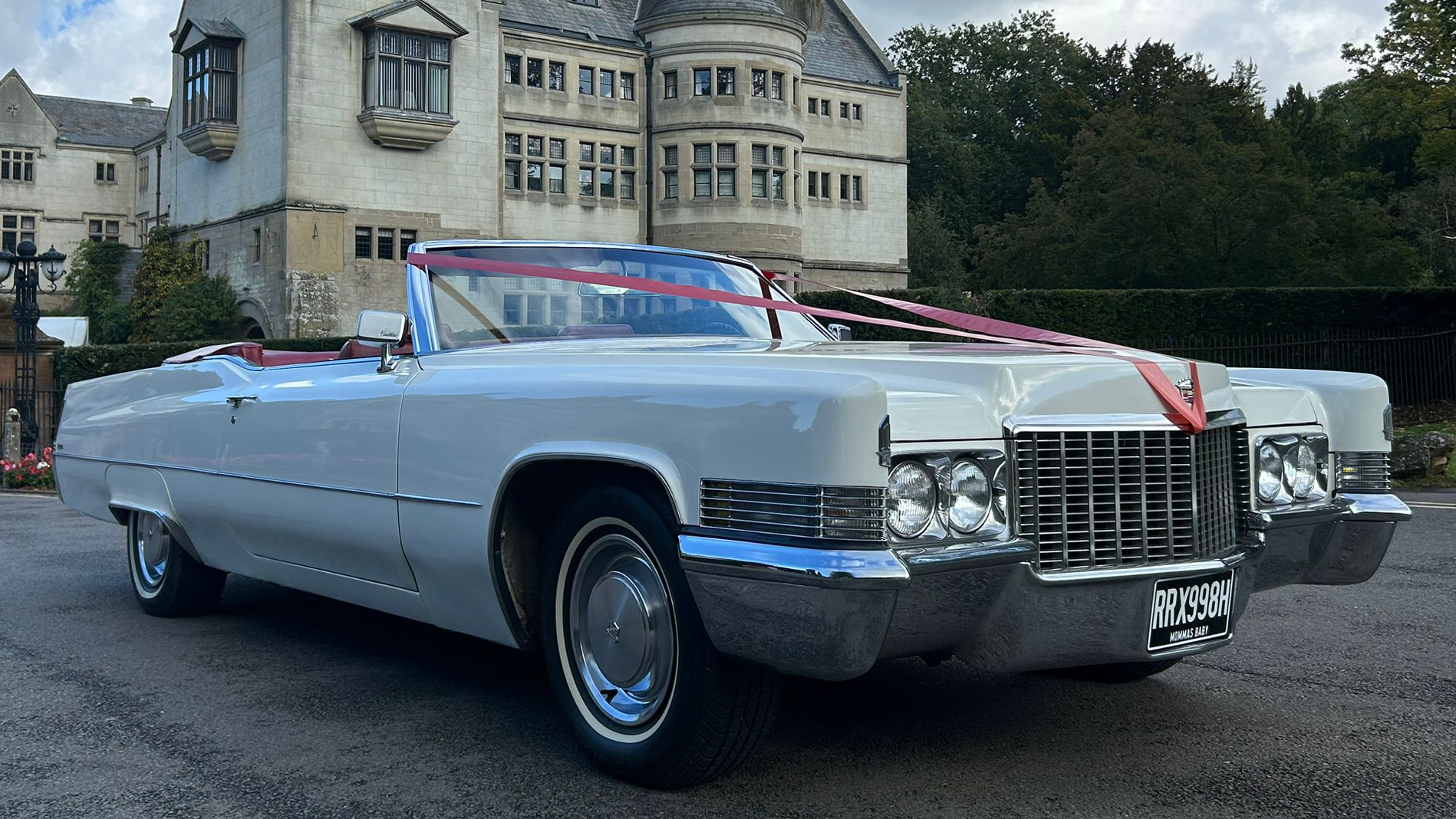 White 1970 Cadillac Coupe DeVille convertible with red leather interior and wedding ribbons on the bonnet, parked outside a stately venue.