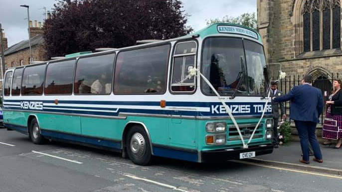 Side view of a turquoise and cream vintage bus parked outside a traditional stone church in Yorkshire with wedding guests nearby.